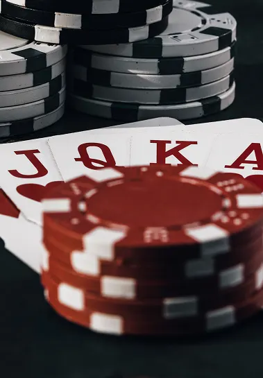 Купи червоних і чорних покерних фішок лежать на темній поверхні, з картами на видимості. Видимі карти - валет, дама, король і туз, що вказує на сильну руку. Сцена випромінює напружену і конкурентну атмосферу азартних ігор.